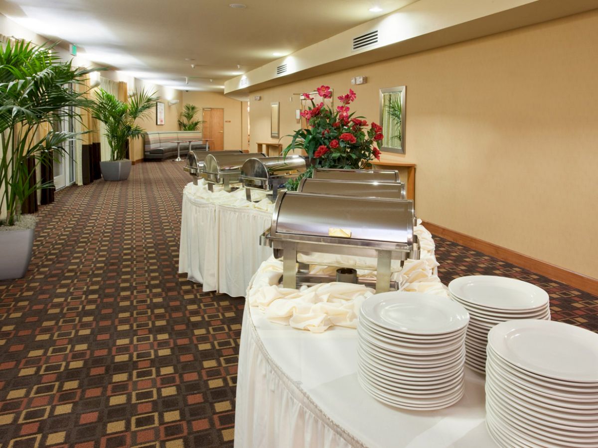 A buffet setup in a carpeted hallway with chafing dishes, stacked plates, and floral arrangements, adjacent to potted plants and a long corridor.